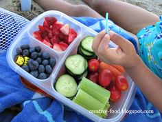 a child holding a plastic container filled with fruit and veggies on the beach
