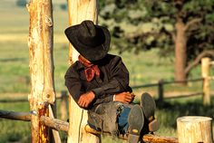 a young boy wearing a cowboy hat sitting on a wooden fence with his skateboard