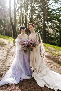 two women in dresses standing next to each other on a dirt road with trees behind them