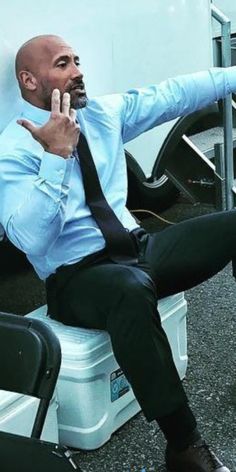 a man in a blue shirt and tie sitting on a white chair with his hand up