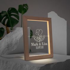 a light up frame with a couple holding hands in the shape of a heart, on top of a table next to a potted plant