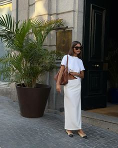 a woman is standing in front of a building wearing a white dress and carrying a brown bag