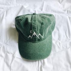 a green hat sitting on top of a white bed covered in sheets and pillows with mountains embroidered on the front