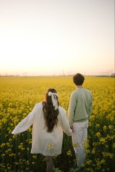 a man and woman standing in a field with yellow flowers looking at each other while the sun is setting