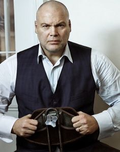 a bald man in a black vest and white shirt sitting on a chair with his hands behind his back