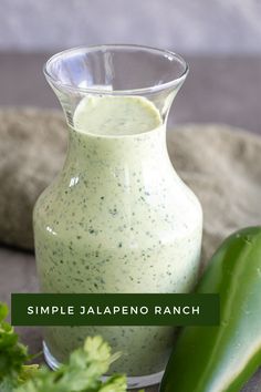 a glass pitcher filled with green sauce next to a cucumber on a table