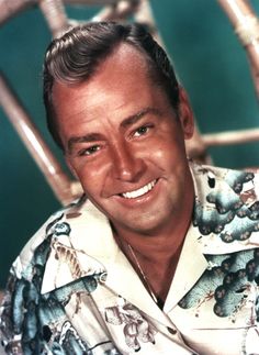 a man in a hawaiian shirt smiles for the camera while sitting on a wicker chair