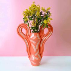 an orange vase with yellow flowers in it on a white table next to a pink wall