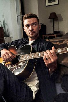 a man sitting on a couch holding an acoustic guitar in his right hand and looking at the camera