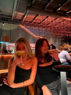 two beautiful women sitting at a table with wine glasses in their hands and one holding a drink