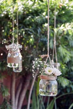 three mason jars filled with white flowers hanging from twine strings in front of trees
