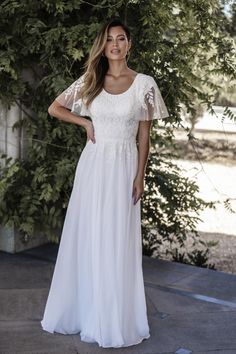 a woman wearing a white dress standing in front of some trees and greenery with her hands on her hips