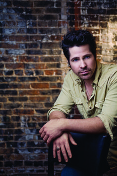 a man sitting in a chair with his arms crossed and looking at the camera while wearing a tan shirt