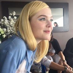 a woman with blonde hair and blue eyes sitting at a table in front of flowers