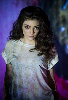 a woman with long hair and blue eyes standing in front of a purple background wearing a white t - shirt