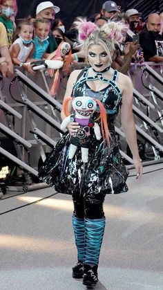 a woman walking down a runway with lots of people behind her wearing costumes and makeup