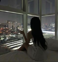 a woman sitting on top of a bed in front of a window looking out at the city