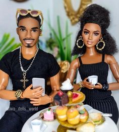 two dolls sitting next to each other on a table with food and drinks in front of them