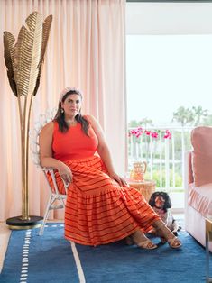 a woman in an orange dress sitting on a chair next to a dog and pink curtains