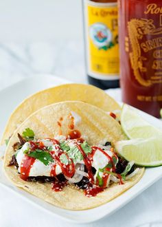 two tacos on a white plate with limes and ketchup in the background