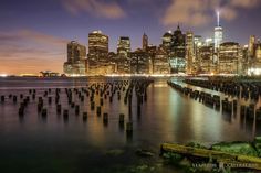 the city skyline is lit up at night as seen from across the water with old docks in foreground