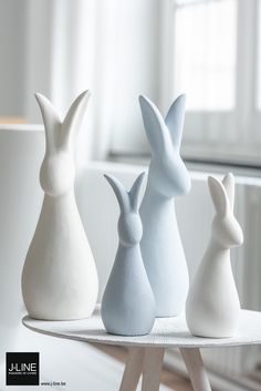 three white and one blue ceramic rabbits sitting on a table in front of a window