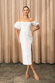 a woman standing in front of a curtain wearing a white dress with an off the shoulder slit