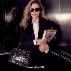 a woman in black jacket and sunglasses holding a book while standing next to an elevator