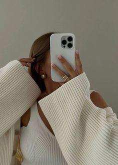 a woman taking a selfie with her cell phone while wearing a white sweater and gold earrings