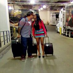 a man and woman kissing while holding luggage in an airport parking garage with semi trucks behind them