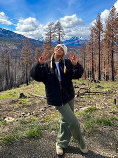 a woman standing in the middle of a forest with her hands up and two fingers out