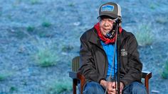 an old man sitting in a chair with a microphone on his lap and wearing a hat