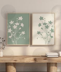 two framed art prints on the wall above a wooden table with vases and books