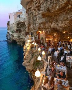 people are sitting at tables on the cliff by the water and eating food in front of them