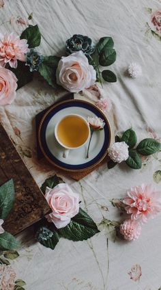 a cup of tea sits on a plate surrounded by pink flowers and greenery, next to an old book
