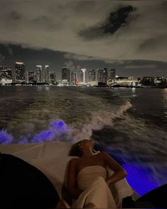 a woman laying on top of a boat in the water at night with city lights behind her