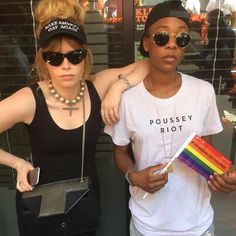 two women standing next to each other holding a rainbow flag and a small black purse