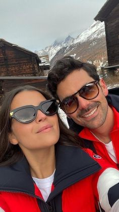 a man and woman taking a selfie in front of a mountain hut with snow covered mountains behind them