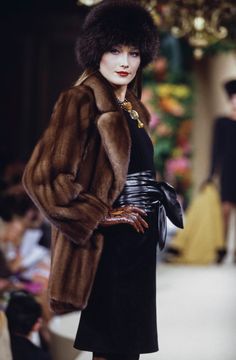 a woman in a fur coat and black dress on the catwalk at a fashion show
