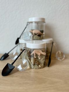 two jars filled with small plastic dinosaurs on top of a wooden table next to utensils