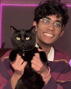 a man holding a black cat in front of a purple wall with lights on it