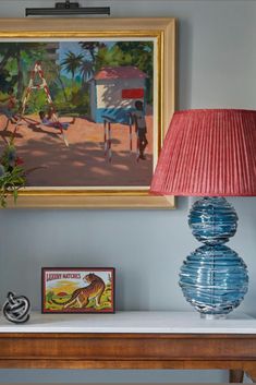 a painting and lamp on a table in front of a blue wall with a red lampshade