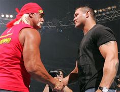 two men shaking hands in front of an audience at a wrestling match, one wearing a red shirt and the other black