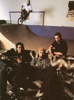 three men are sitting on the floor in front of a skateboard ramp and one man is doing a trick