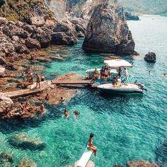 people are swimming in the clear blue water near some rocks and boats with umbrellas