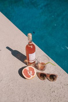 a bottle of wine, sunglasses and grapefruit next to a pool with water