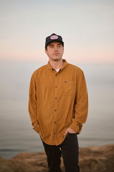 a man standing on top of a cliff near the ocean wearing a brown shirt and black hat