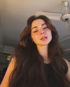 a woman with long brown hair standing in front of a ceiling fan and looking at the camera