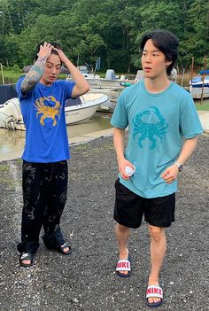 two young men standing next to each other in front of boats on the water with trees in the background