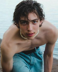 a shirtless young man posing on the beach with his surfboard and pearls around his neck